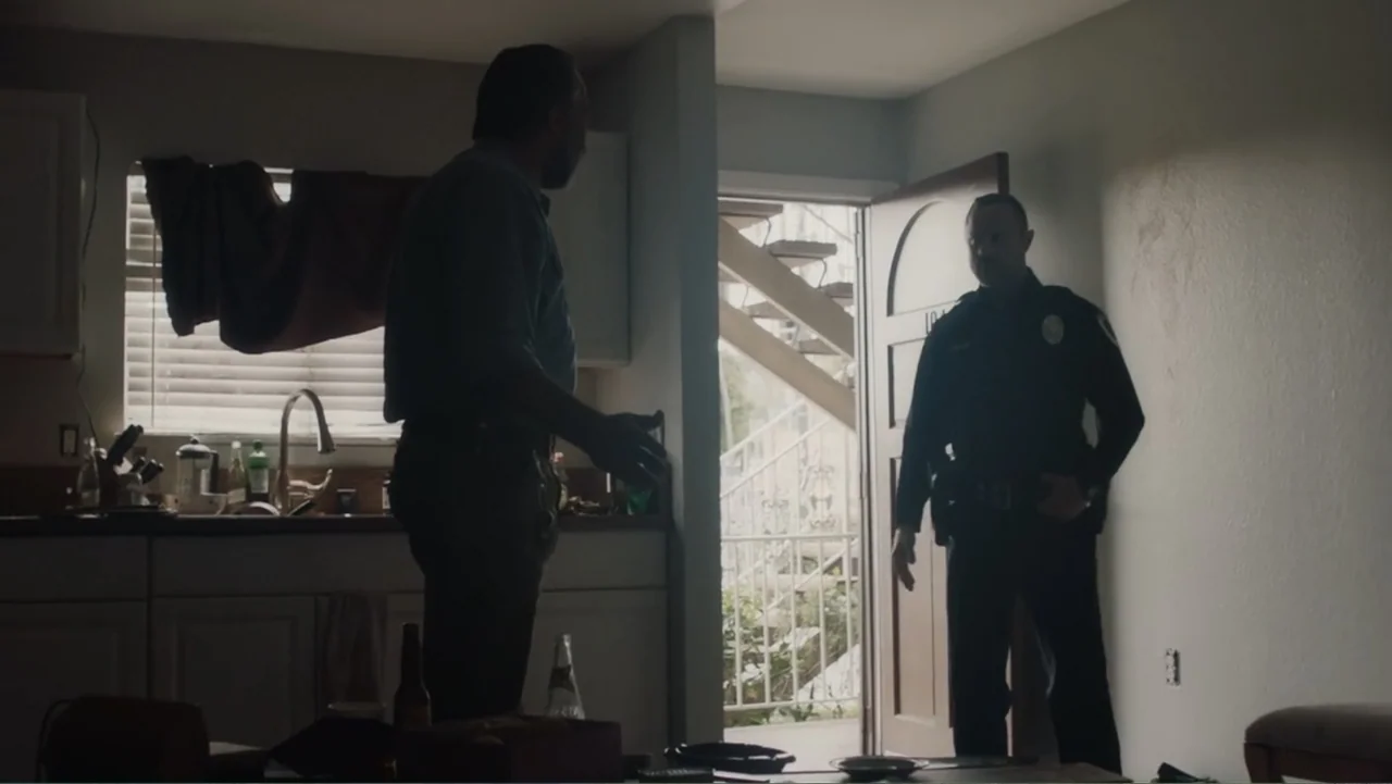 The Cleaner (2021) movie still: medium — A dimly lit room with a kitchen area and an open doorway leading outside. A man in civilian clothes faces a…