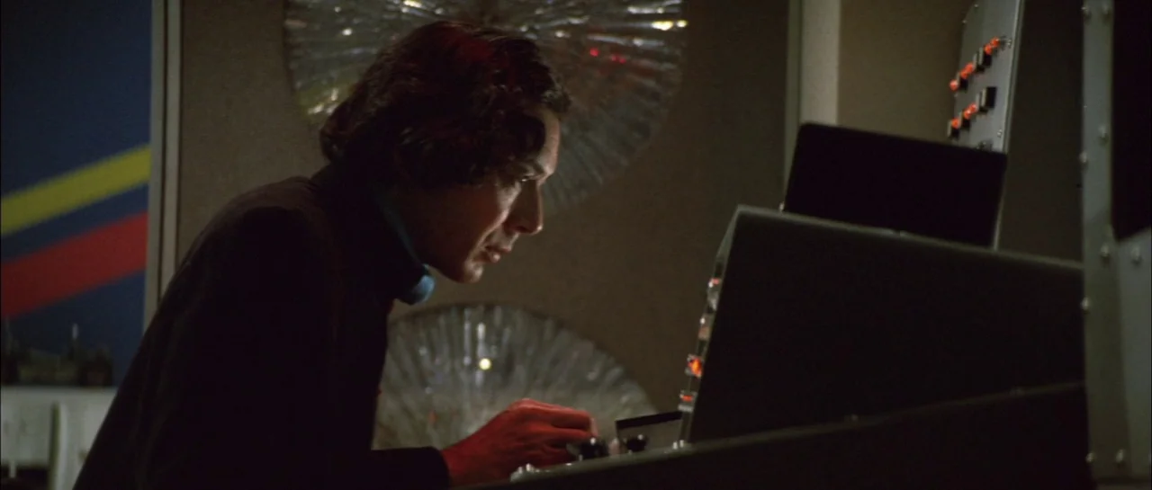 Godzilla Vs Megalon (1973) movie still: medium — A dimly lit control station features a complex console with glowing red buttons. A man in dark…