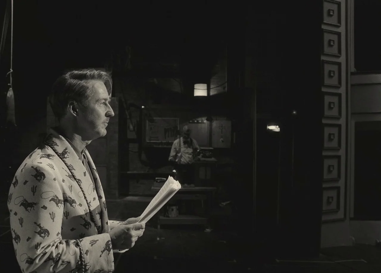 Asteroid City (2023) movie still: medium — A dimly lit theatrical stage setting with a blurred figure in the background. A man in a patterned suit…