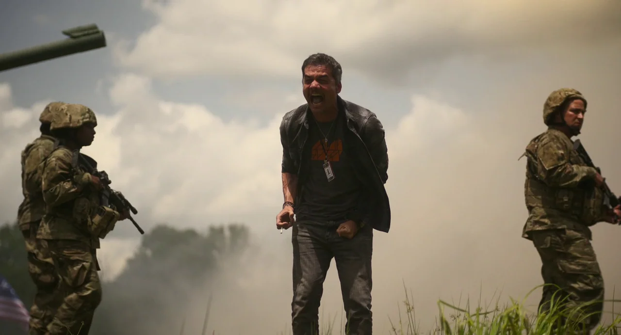Civil War (2024) movie still: fullbody — An open grassy field under a cloudy sky during a military deployment. A man stands between two armed…