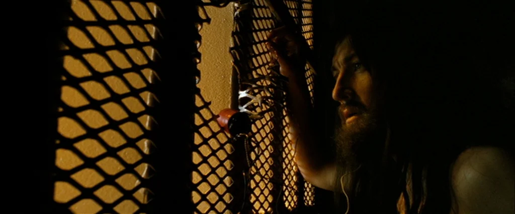 Hunger (2008) movie still: medium — A man with long hair and a beard peers through a heavy metal grate. The enclosure creates a grid pattern of…
