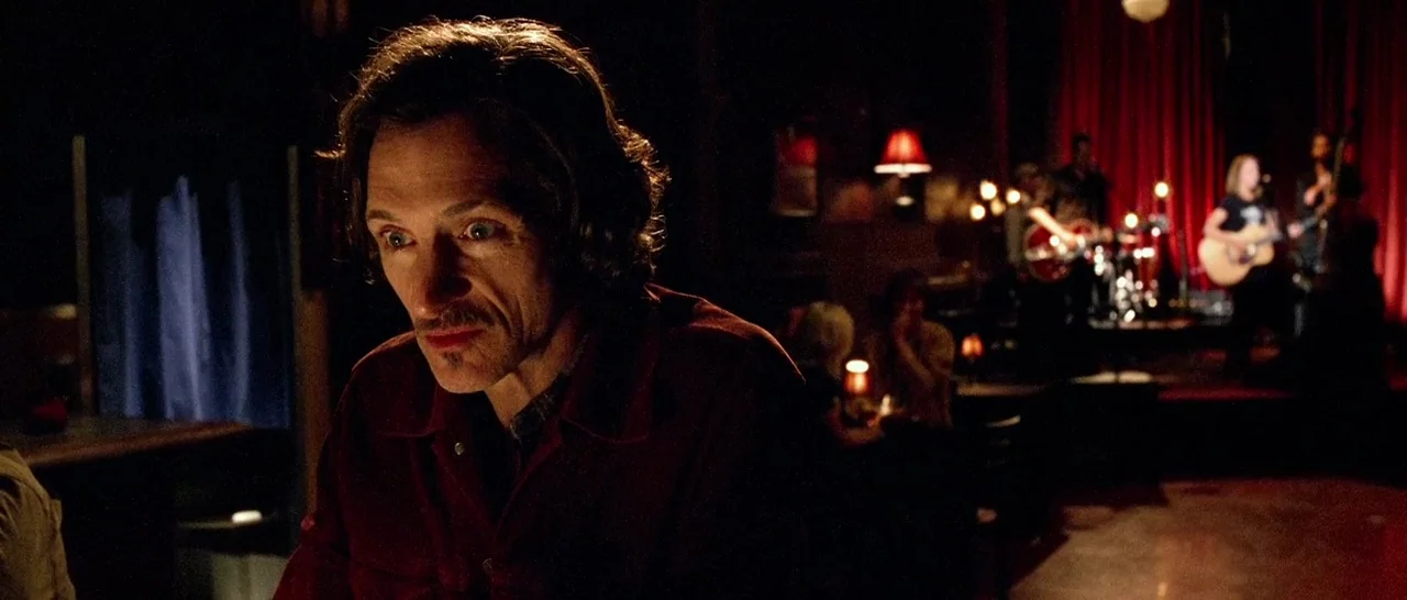Too Late (2015) movie still: medium — A dimly lit room with red curtains and a performance stage in the back. A man with dark hair looks toward the…