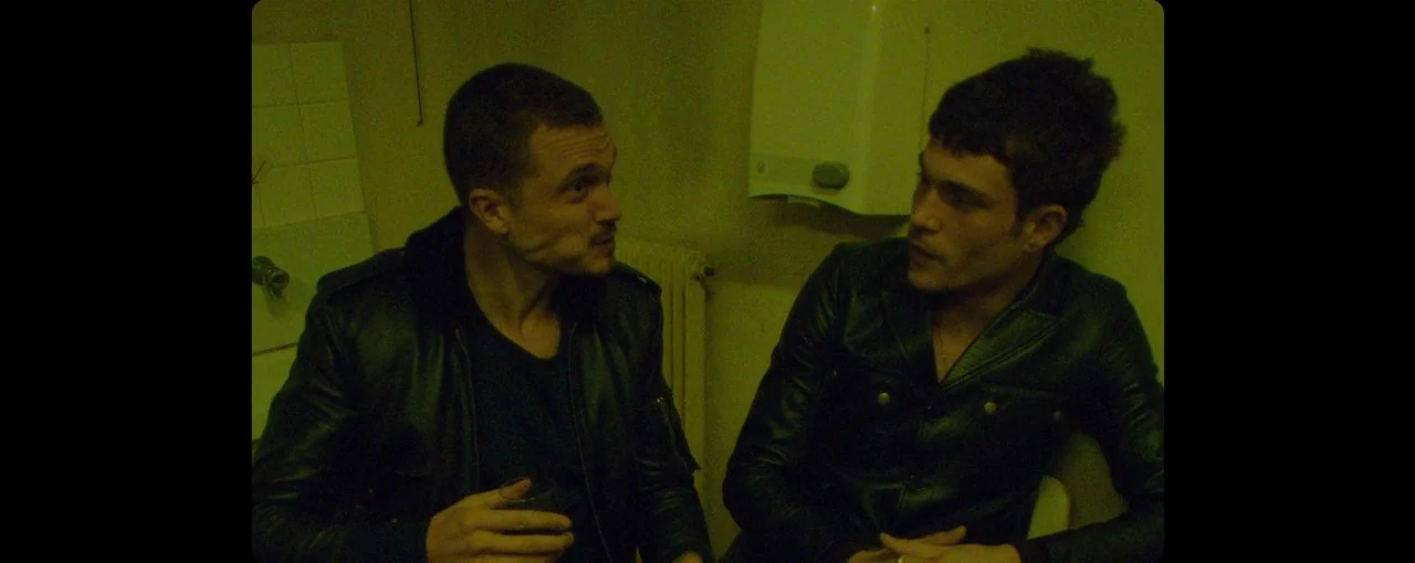 Lux Æterna (2019) movie still: medium — Two men sit in a dimly lit, green-hued restroom interior with tiled walls. They are positioned closely,…