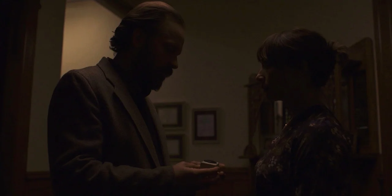 The Sound of Silence movie still: medium — A dimly lit residential hallway with framed artwork hanging on a dark wall. A man and a woman stand face…