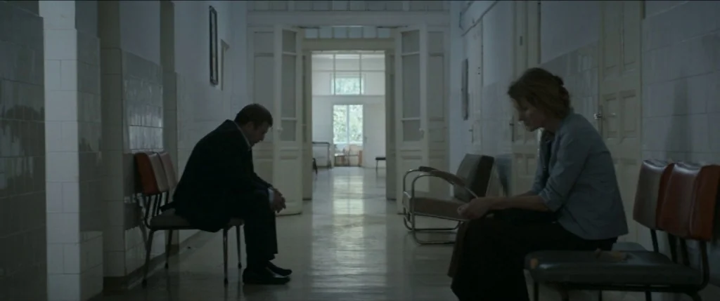 Graduation (2016) movie still: wide — A long, clinical hospital hallway with light-colored tiled walls and linoleum flooring. A man and woman sit on…