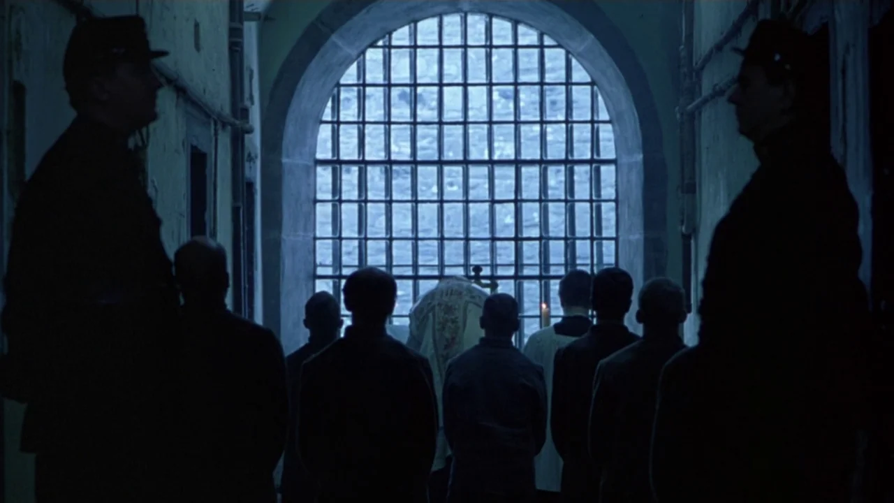 Michael Collins (1996) movie still: wide — A group of men stand in a dark, narrow corridor facing an arched window. The window is covered with iron…