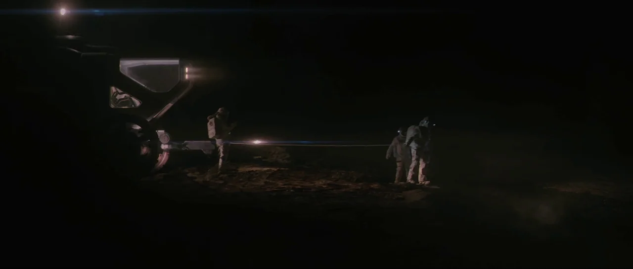 The Last Days On Mars (2013) movie still: wide — A dark, rocky lunar landscape is illuminated by the bright headlights of a vehicle. Three figures…