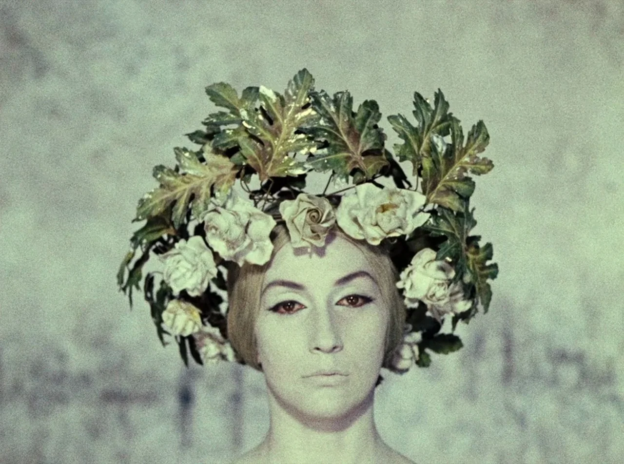 The Colour of Pomegranates (1969) movie still: closeup — A woman wears a large floral crown against a plain, light-colored backdrop. The woman…