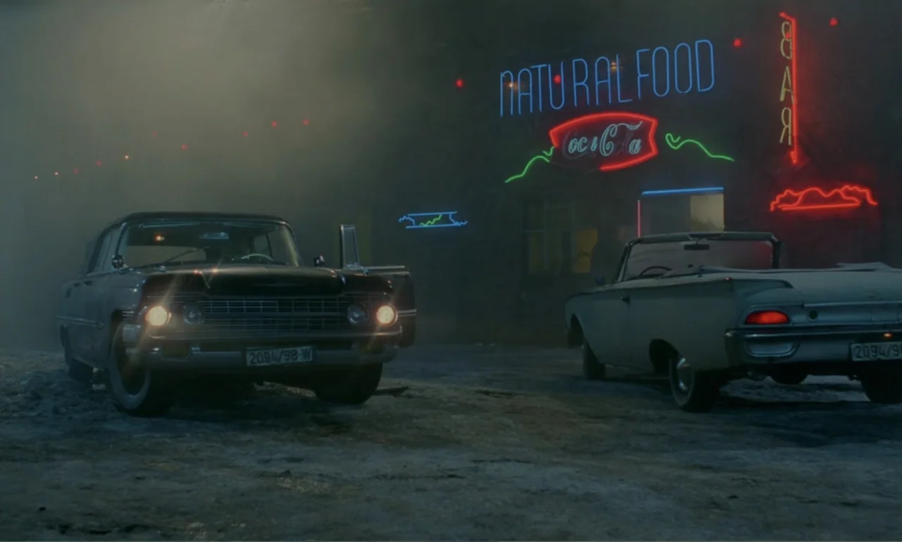 Ga-ga: Glory to the Heroes (1986) movie still: wide — A dark, foggy exterior of a roadside diner at night with vibrant neon signage. Two vintage…