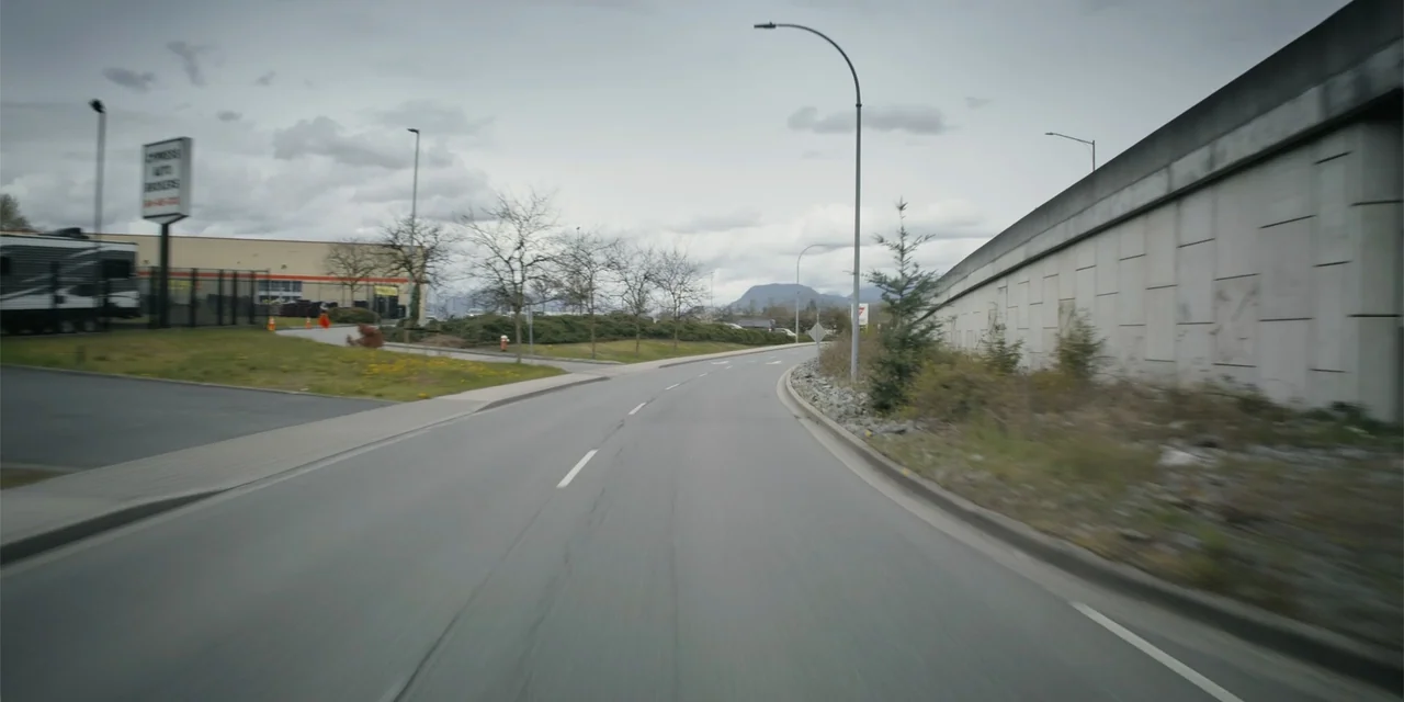 Playdate (2025) movie still: establishing — An asphalt road curves past a concrete wall and commercial buildings under grey skies. Sparse vegetation…