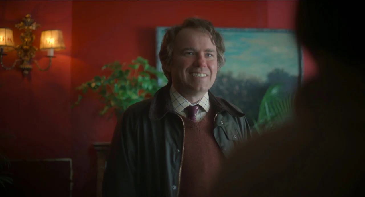 Men (2022) movie still: medium — A man stands before a bold red wall adorned with an ornate wall sconce. He is wearing layered outerwear and smiles…