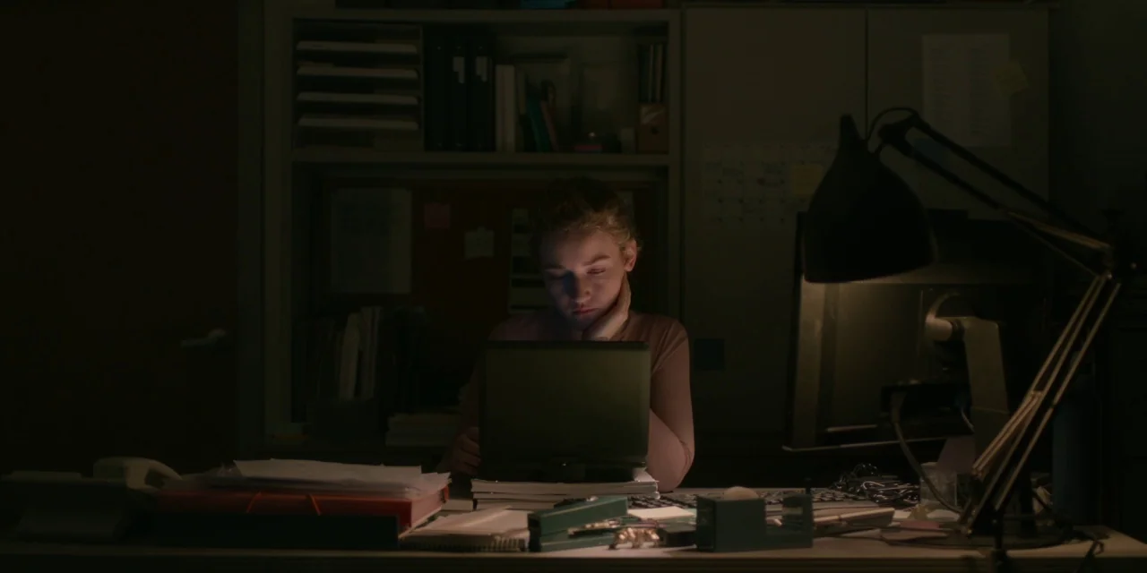 The Assistant (2019) movie still: medium — A dimly lit office workspace contains cluttered paperwork, a desk lamp, and storage cabinets. A young…