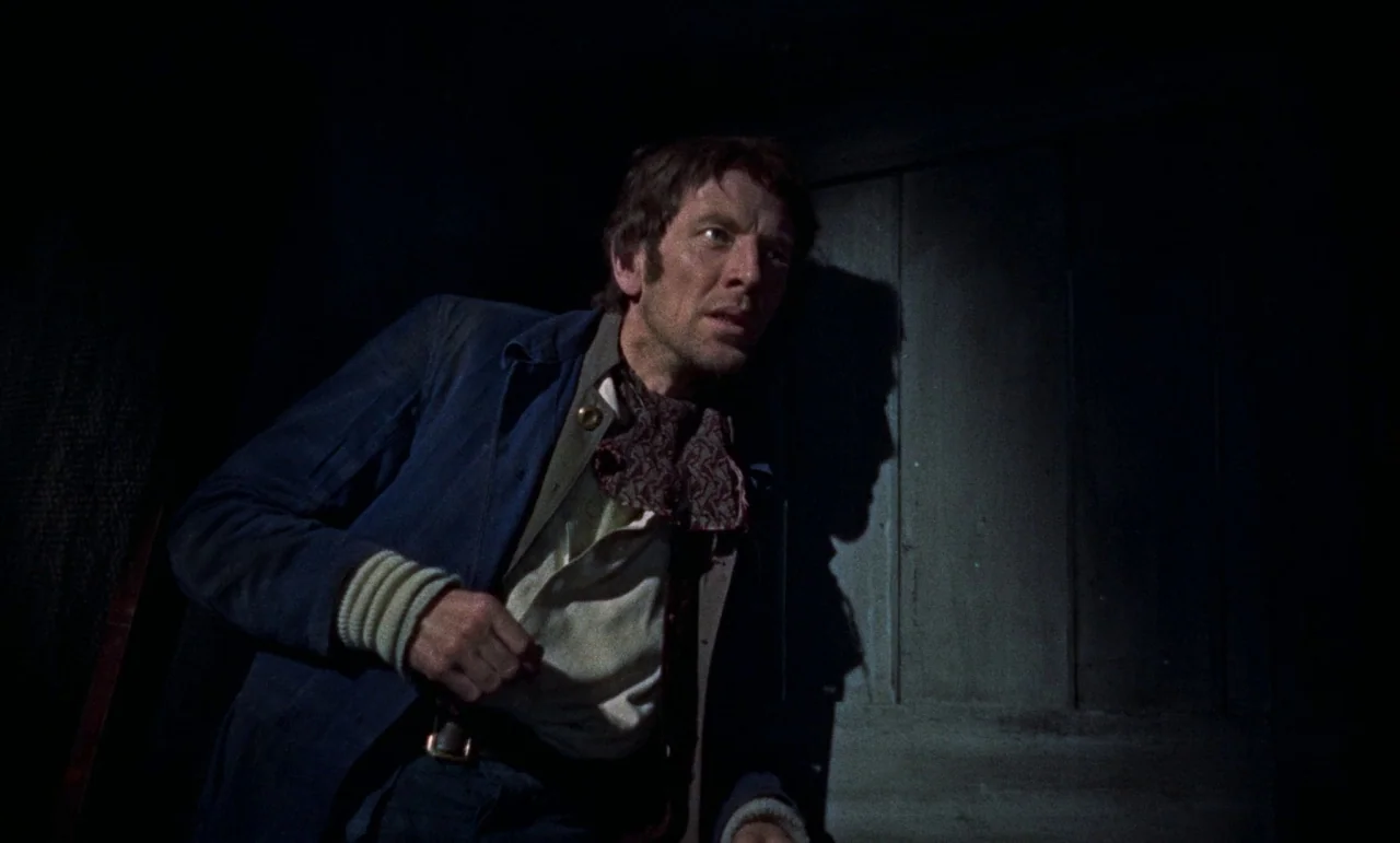 The Revenge of Frankenstein movie still: medium — A dark, dimly lit room with simple vertical wooden planks forming the wall surface. A man stands…