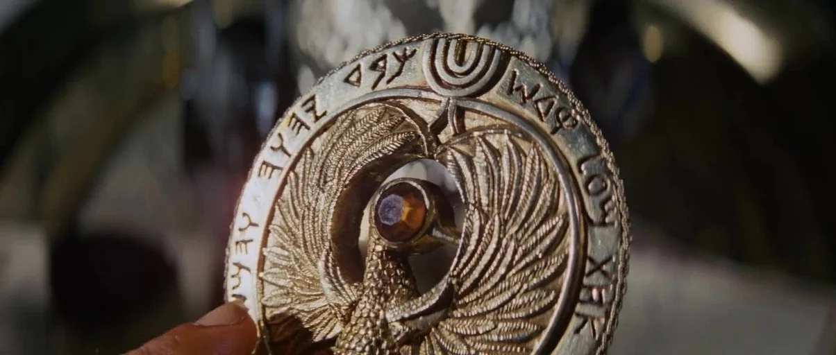 Raiders Of The Lost Ark (1981) movie still: closeup — A close-up shot of an intricate gold medallion featuring a central amber gemstone. The outer…
