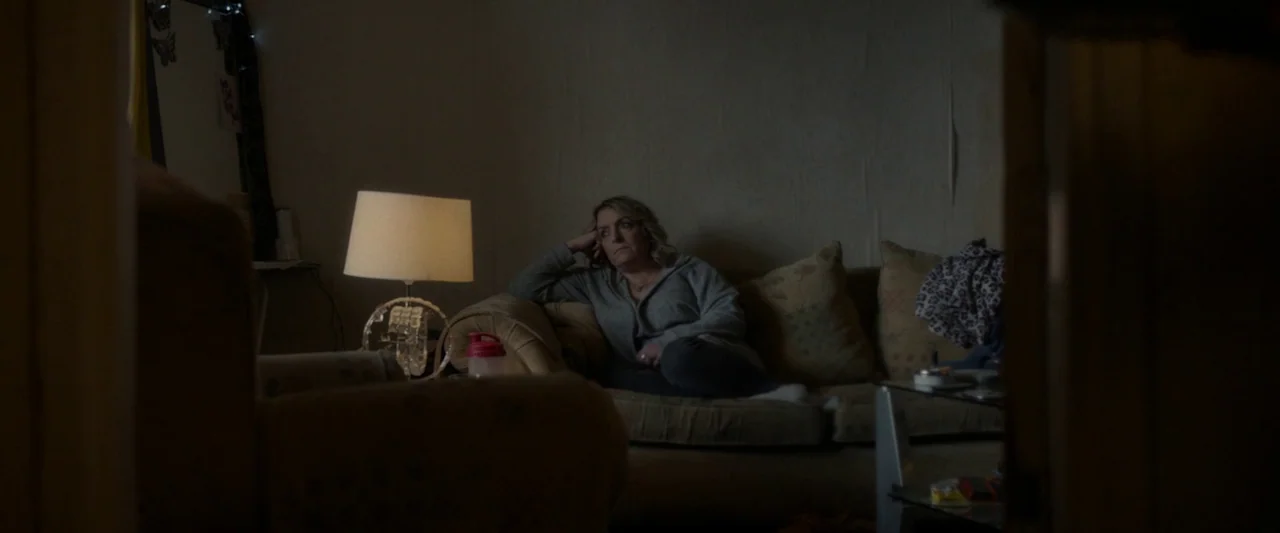 Ali & Ava (2021) movie still: medium — A dimly lit room features a worn beige sofa against a plain wall. A middle-aged woman sits on the sofa with…