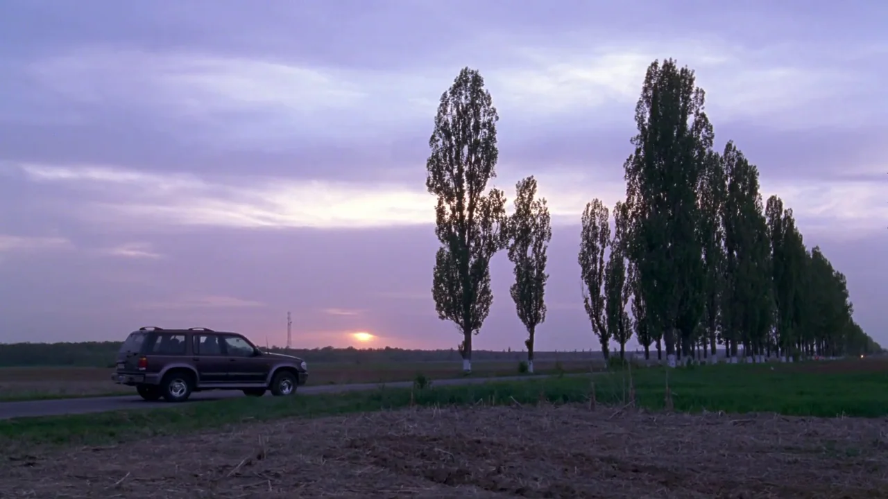 Hellraiser: Hellworld (2005) movie still: wide — A dark SUV sits parked on a narrow asphalt road next to a tilled field. A row of tall, slender…