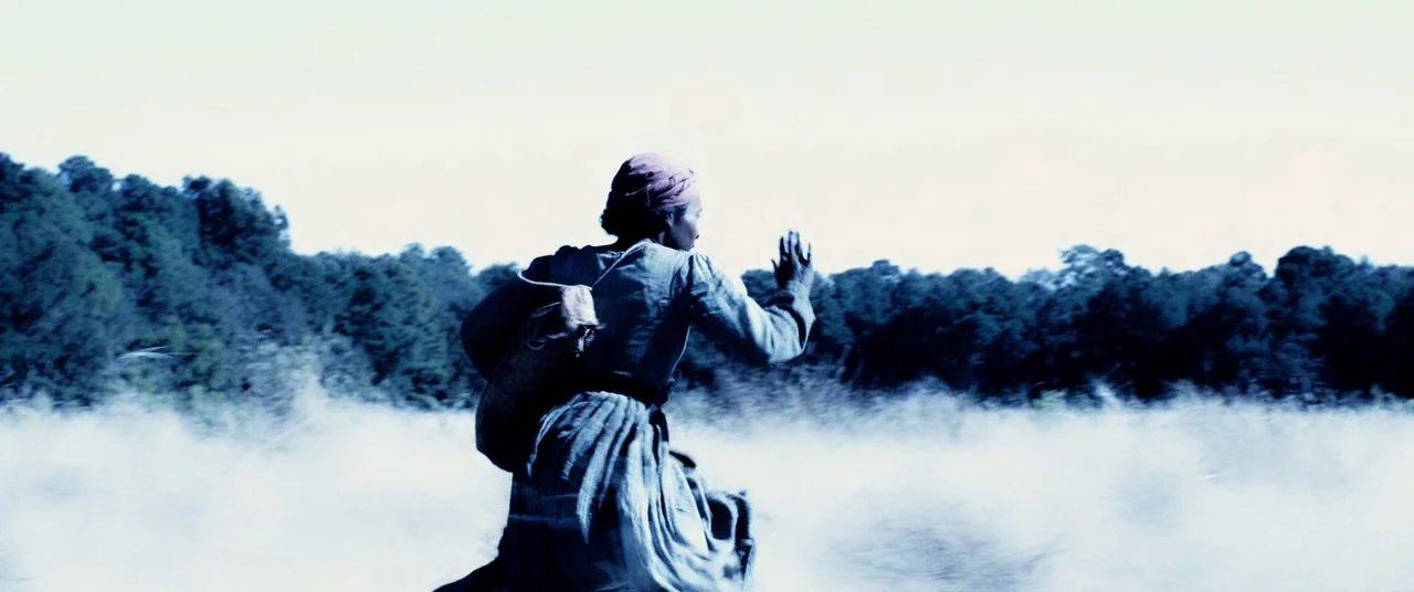 Harriet movie still: fullbody — A woman runs through a misty field beneath an overcast sky with distant trees. She travels from left to right while…