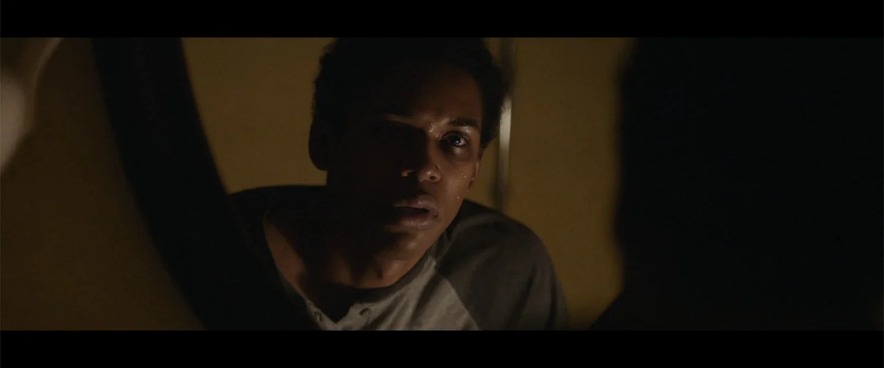 It Comes at Night (2017) movie still: closeup — A dimly lit room with dark walls and shadows obscuring the background. A young man stares intently…