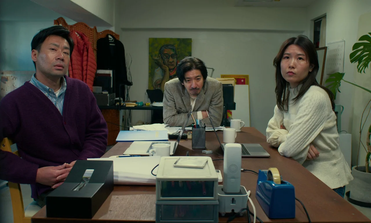 Evil Does Not Exist (2023) movie still: medium — A cluttered communal work table in a bright, white-walled room contains laptops and stationery.…