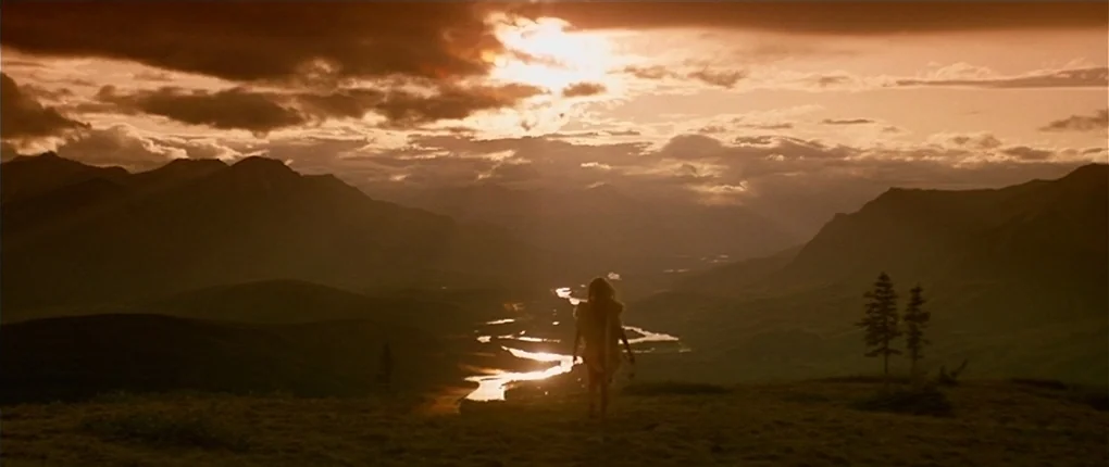 Clan of the Cave Bear (1986) movie still: wide — A vast mountain landscape with a winding river valley under a sunset sky. A solitary woman walks…