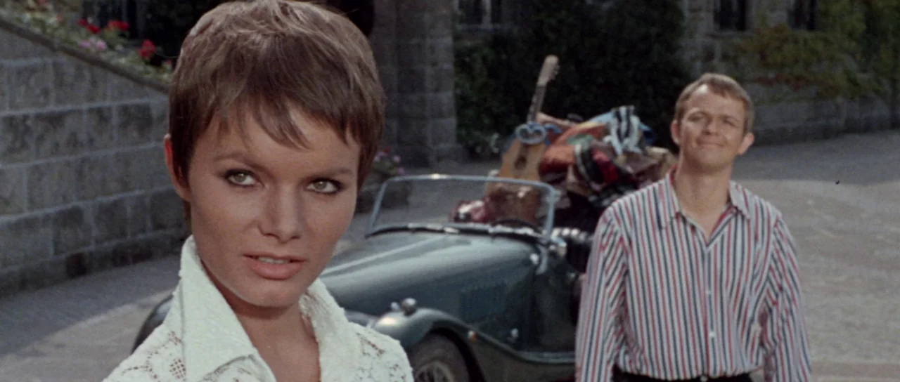 Orgasmo (1969) movie still: medium — A stone paved area sits in front of a building with a dark car parked. A woman looks toward the camera with a…