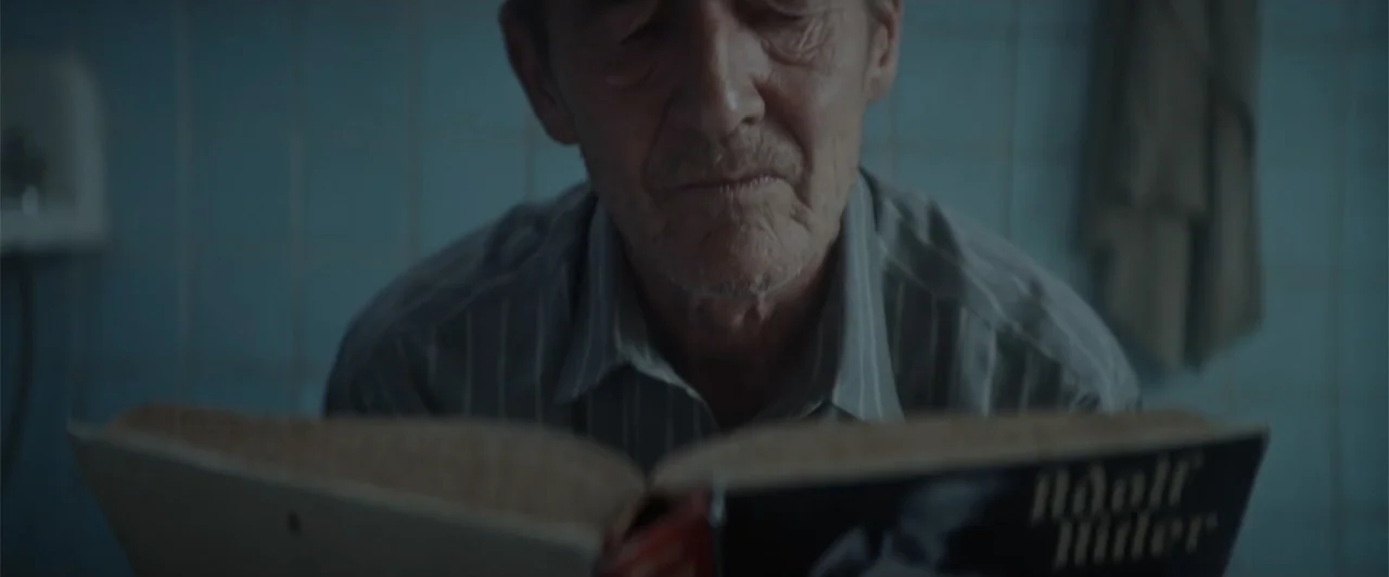 My Neighbor Adolf (2022) movie still: medium — An interior space with blue-tiled walls is illuminated by dim, soft light. An older man sits alone…