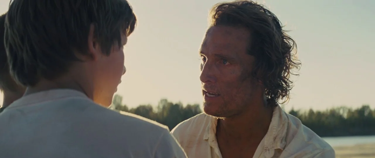 Mud (2012) movie still: medium — A riverside at sunset with trees visible in the soft-focus background. A man with a dirty face speaks directly to a…