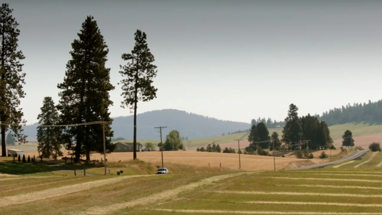 At Middleton (2013) movie still: wide — An expansive rural landscape features dry golden fields and scattered tall coniferous trees. A small car…
