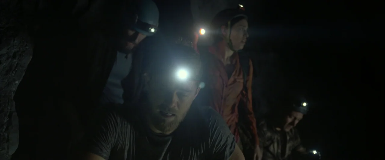 Black Water: Abyss (2020) movie still: medium — A group of explorers traverses a narrow rocky cave system in deep darkness. The subjects wear safety…