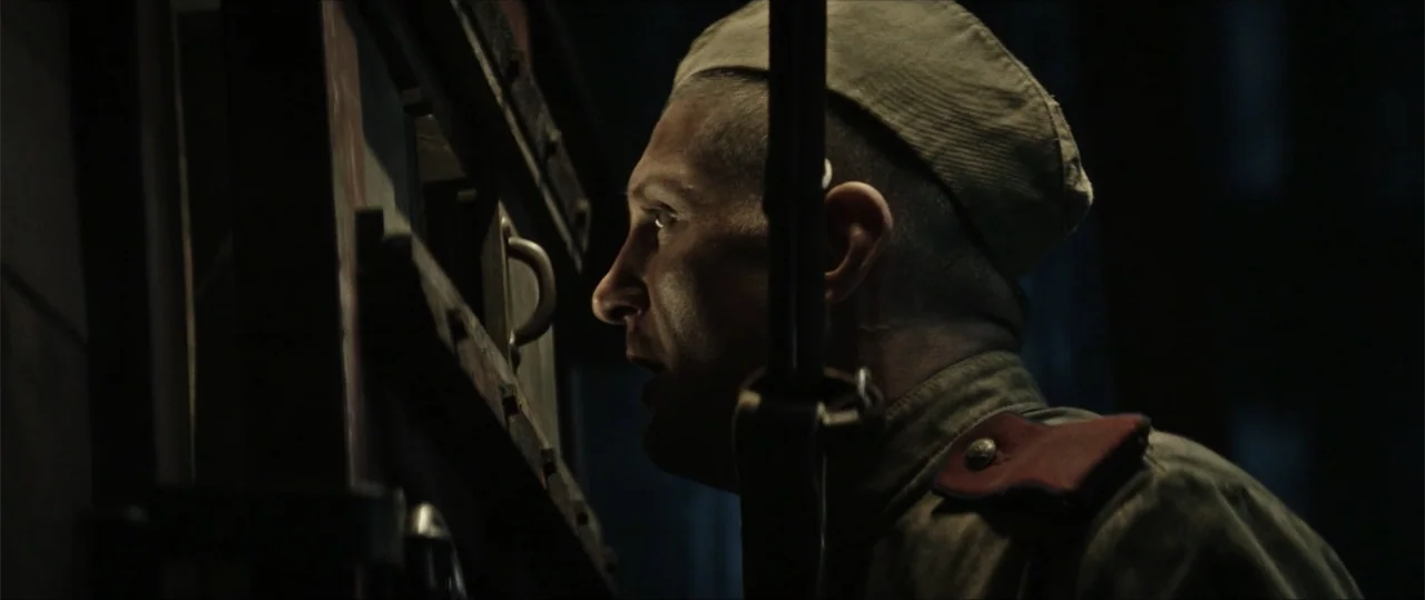 Sisu: Road to Revenge (2025) movie still: closeup — A dimly lit interior space with a thick, bolted wooden door and dark shadows. A soldier in…