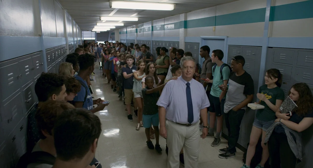 Eighth Grade (2018) movie still: fullbody — A long school corridor lined with gray lockers has a teal accent stripe painted horizontally. A man in a…