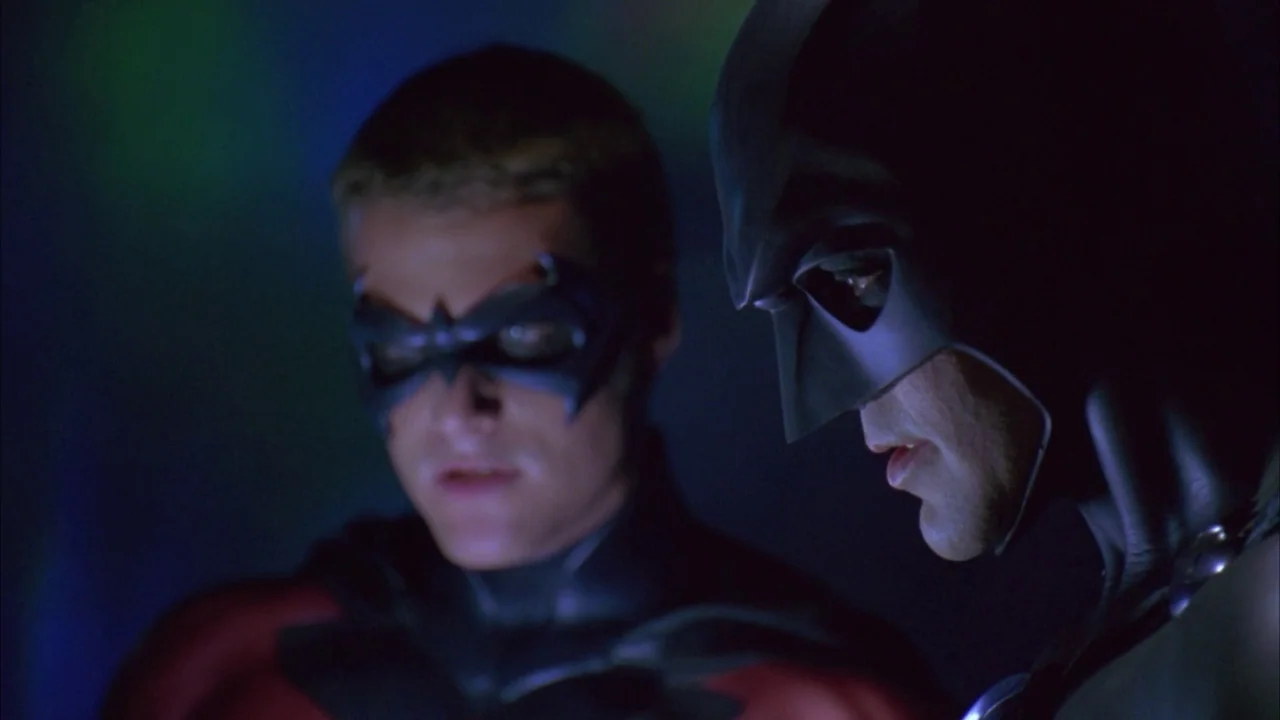 Batman & Robin (1997) movie still: medium — Two figures in superhero costumes occupy a dimly lit, dark space with neon accents. Batman and Robin…