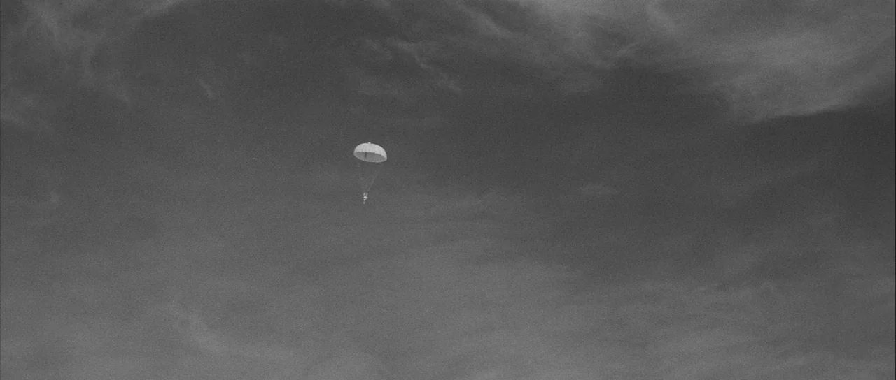 The Tarnished Angels (1957) movie still: establishing — A vast, overcast sky fills the frame with layered grey cloud formations. A lone paratrooper…