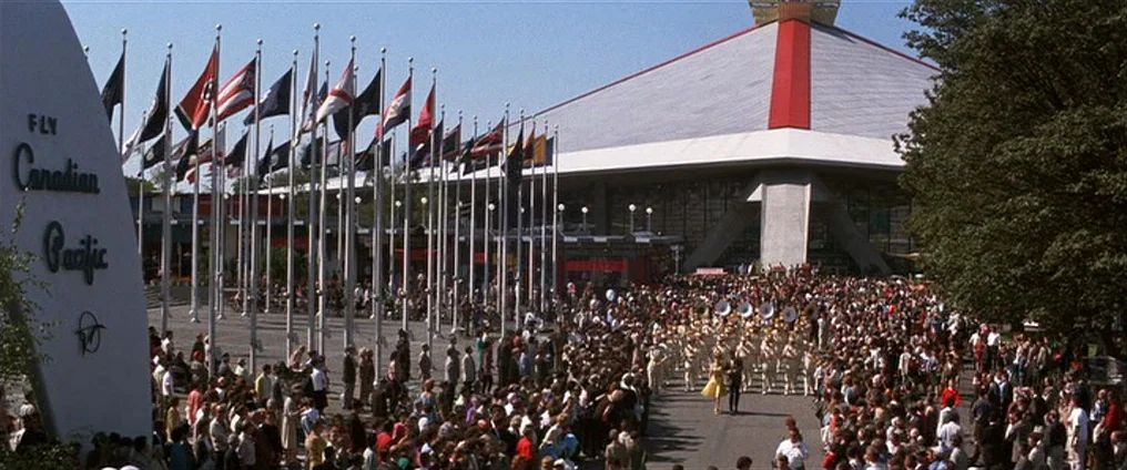 It Happened At The World’s Fair (1963) movie still: wide — A modern geometric pavilion building stands behind a wide plaza with numerous national…