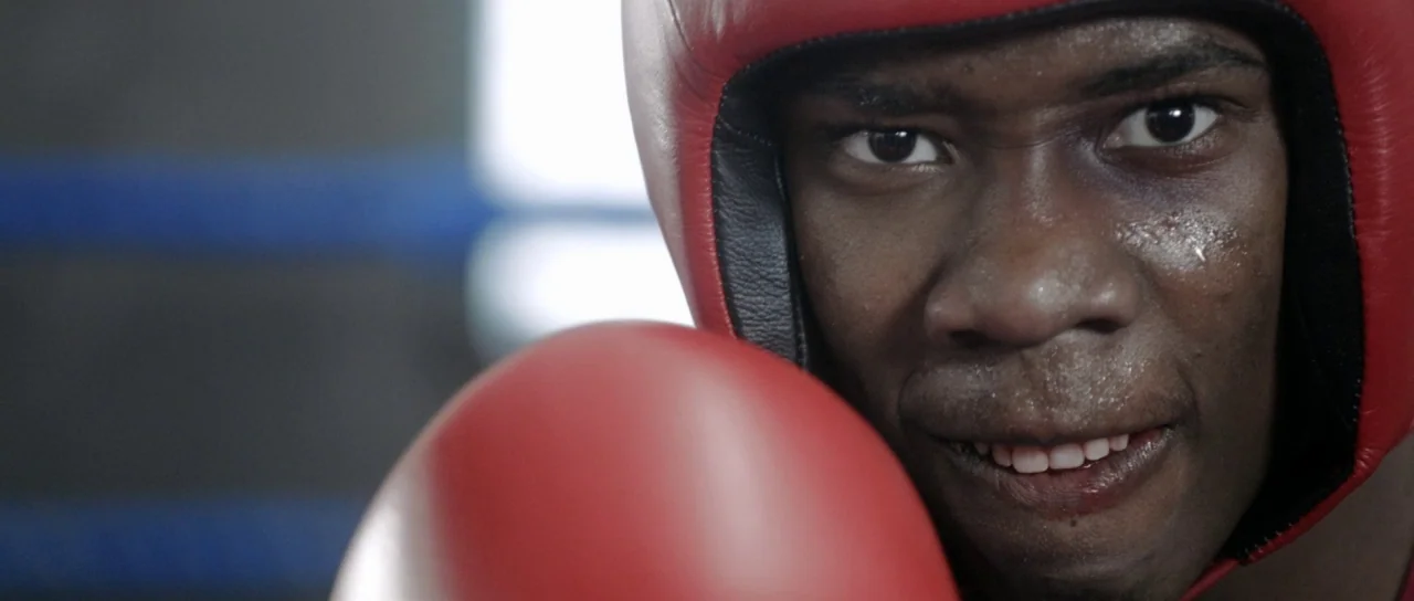 The Fits (2016) movie still: closeup — A boxing ring background with blurred blue and black padding. A man wearing red boxing headgear and a glove…