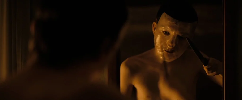 Let Me In (2010) movie still: medium — A dimly lit space features a man viewing his reflection in a framed mirror. The man wears a pale mask and…