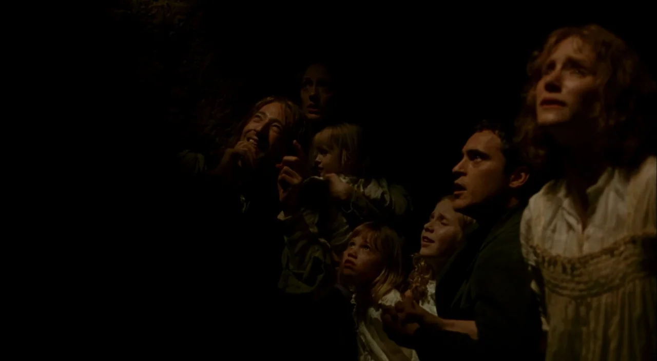 The Village (2004) movie still: medium — A group of several people stand in a dark, confined space with shadowed walls. They are all looking upward…
