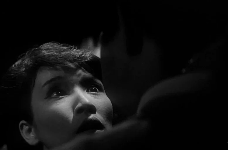 Branded To Kill (1967) movie still: closeup — A dark, nondescript interior space features a woman lit by a sharp, directional light. A dark…