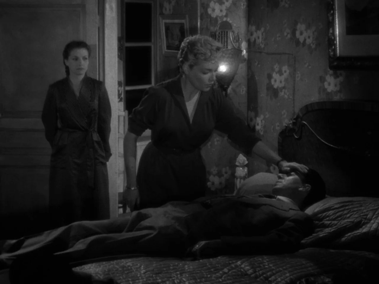 Diabolique (1955) movie still: medium — A dimly lit bedroom with patterned wallpaper and an unmade bed featuring a headboard. A woman leans over a…