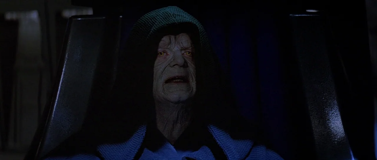 Return Of The Jedi (1983) movie still: closeup — An elderly figure with pale, weathered skin sits in a dark, shadow-drenched environment. He wears a…