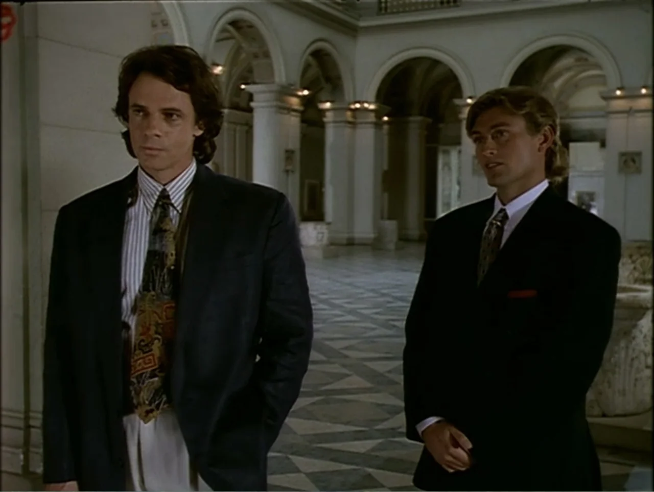 A Change of Place (1994) movie still: medium — A grand interior space features classical architectural columns and a geometric tiled floor. Two men…