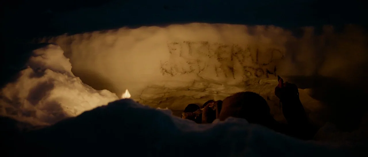 The Revenant (2015) movie still: medium — A dark, cramped snow cave features a wall carved with text written in blood. A faint flame flickers in the…