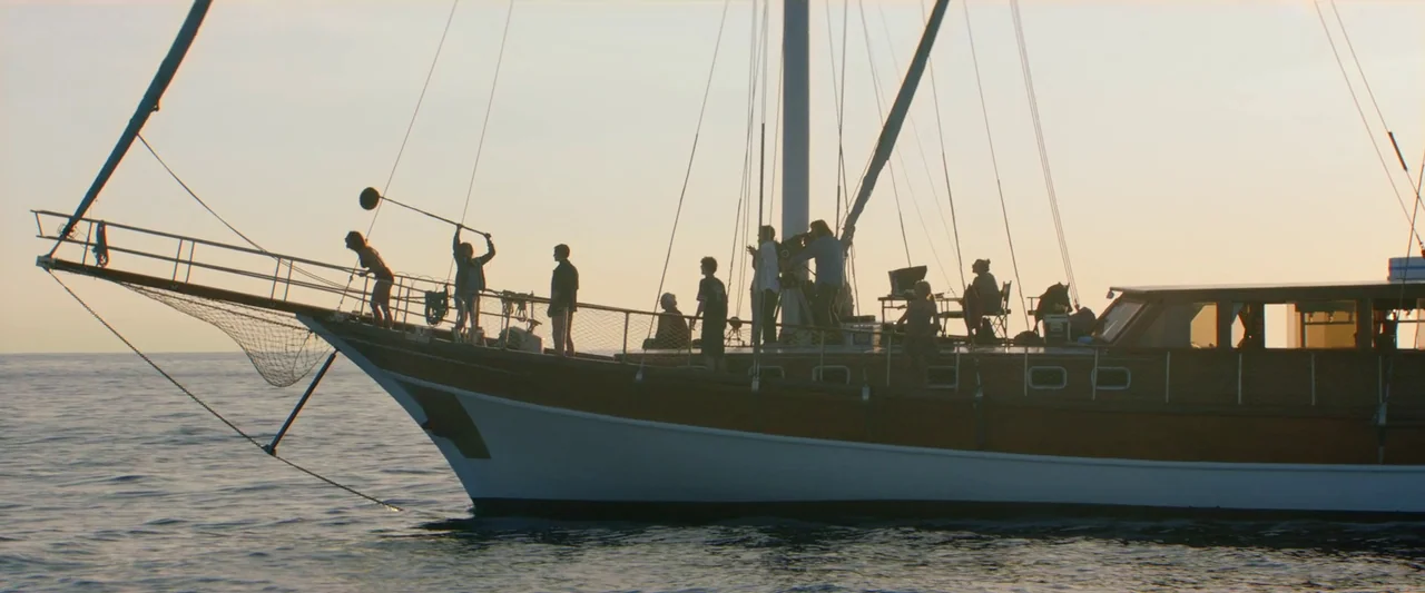 Sibyl (2019) movie still: wide — A film crew works on the deck of a classic wooden sailing yacht at sunset. Several people operate professional…