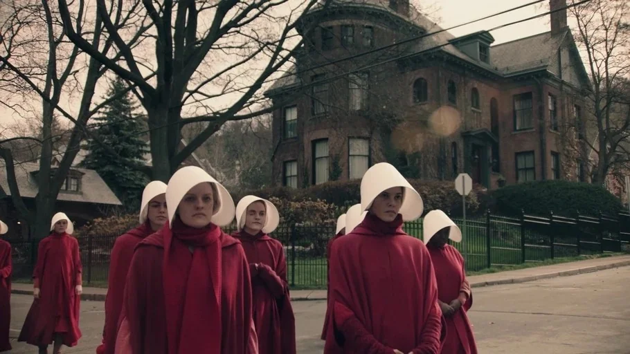 The Handmaid's Tale (2017) movie still: medium — A group of women in red cloaks and white bonnets walk down a quiet street. A large, historic brick…