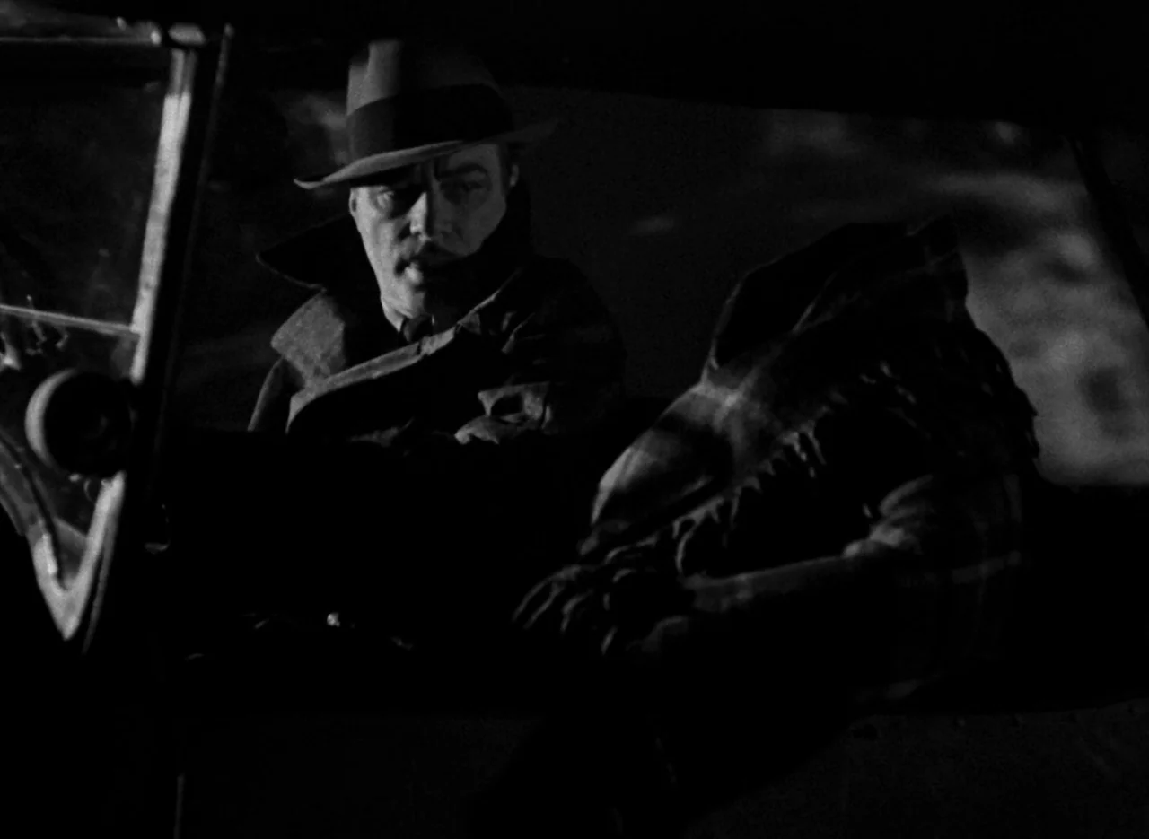 The Invisible Man (1933) (1933) movie still: medium — A man wearing a fedora sits in the driver seat of an automobile at night. He is positioned…