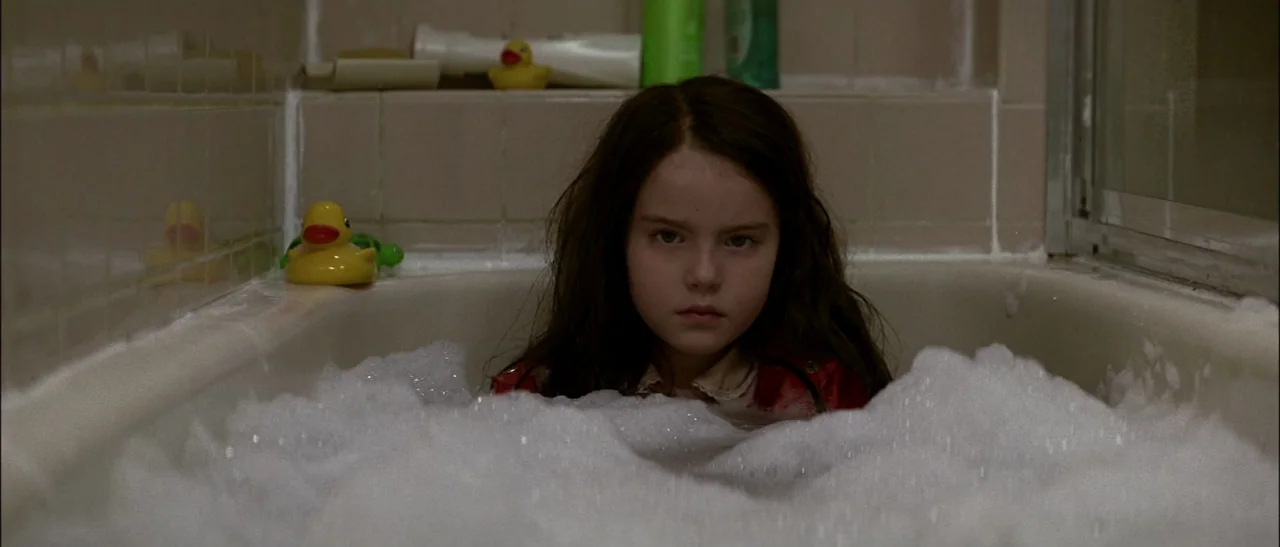 Dark Water (2005) movie still: medium — A child sits in a beige tiled bathtub filled with thick white soap suds. Several yellow plastic rubber ducks…