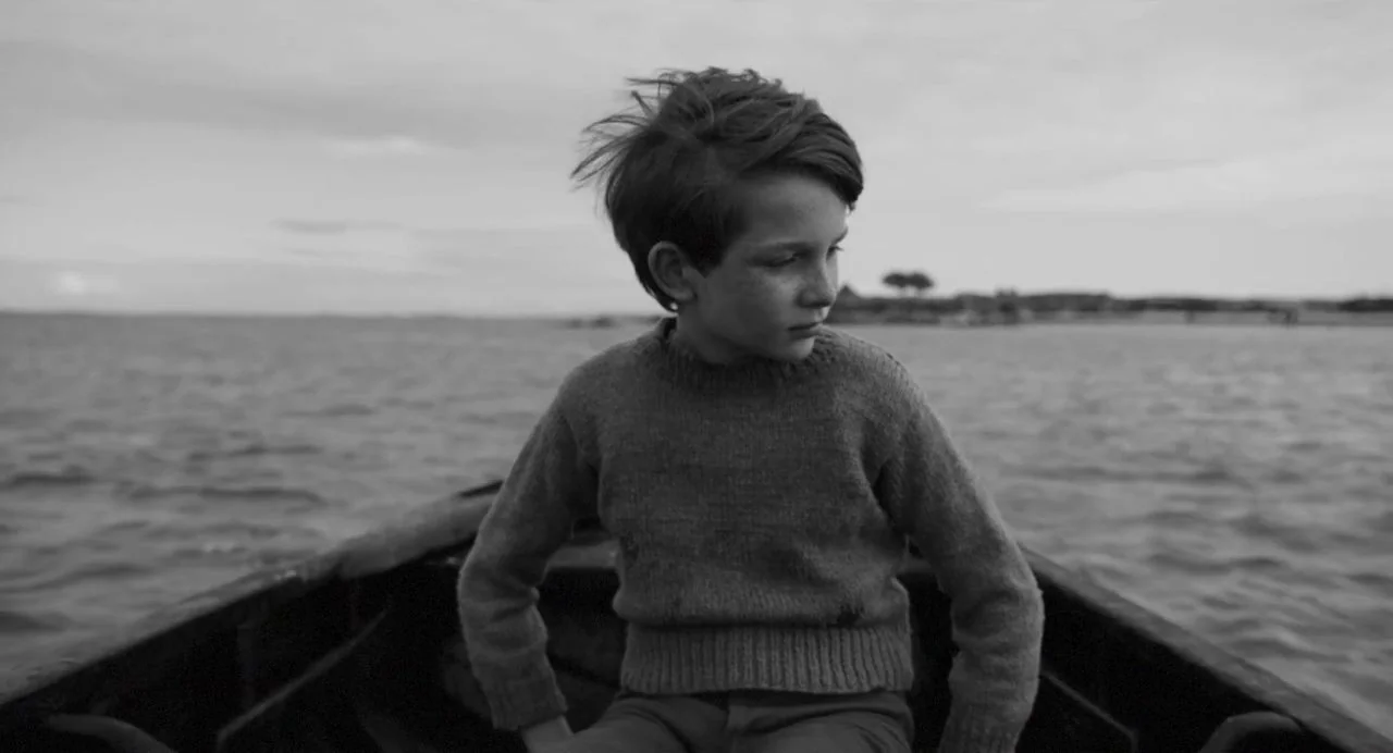 Song of Granite (2017) movie still: medium — A wooden rowboat floats on a wide, calm body of water under a cloudy sky. The boy sits centered in the…