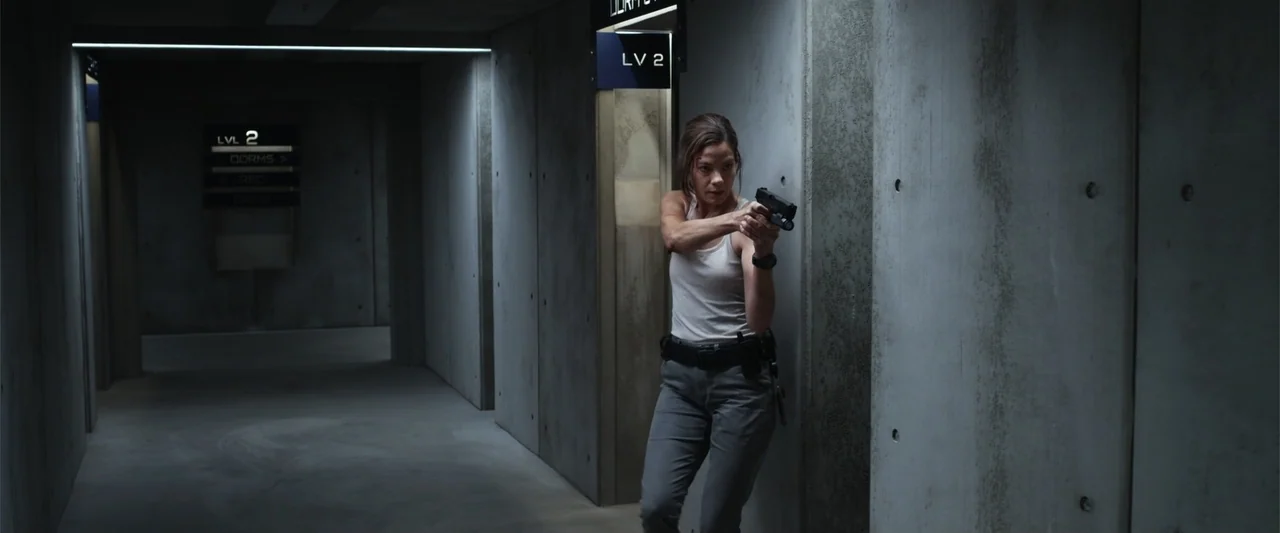Black Site (2022) movie still: medium — A stark industrial corridor features bare concrete walls and metal signage marking levels. A woman stands…
