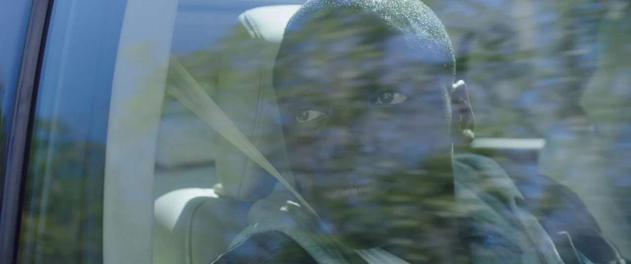 Get Out (2017) movie still: closeup — The interior of a moving car shows a young man seated by the window. Motion blurred trees are visible through…