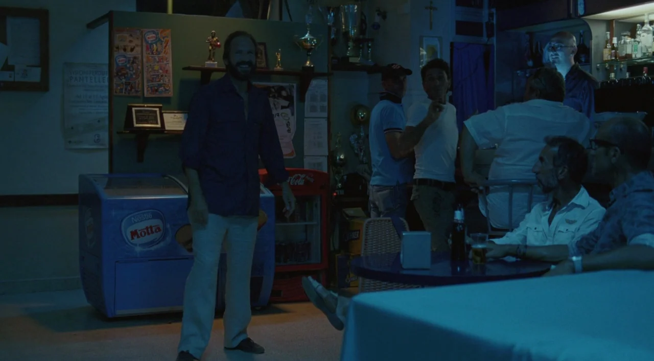 A Bigger Splash (2015) movie still: medium — A dimly lit bar with a blue ice cream freezer in the foreground. Several men are gathered in the…